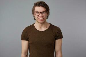 photo de Jeune fou gars avec des lunettes porte dans Vide T-shirt, des stands plus de gris Contexte et en colère regards à le caméra.