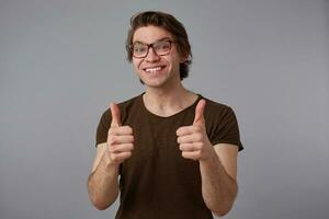 portrait de Jeune de bonne humeur gars avec lunettes, des stands plus de gris Contexte avec content expression, spectacles comme geste et largement sourit. photo