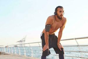 portrait de Jeune sportif barbu homme, du repos après Matin déchiquetage à le bord de mer, Ecoutez préféré mélanger sur écouteurs. photo