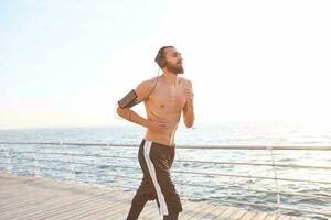 portrait de Jeune Beau sportif gars avec barbe, fonctionnement à le bord de mer, Ecoutez préféré mélanger sur écouteurs. prendre plaisir le Matin et jongler. photo