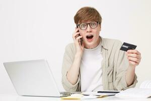 parlant à une banque directeur sur le téléphone, Jeune gars dans des lunettes en portant une crédit carte, un expression de inattendu joie sur le visage de le fait cette il a été possible à résoudre le problème, large ouvert bouche photo