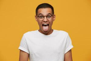 portrait de attrayant Jeune homme dans lunettes, blagues, coincé en dehors le sien langue, grimaces regards drôle, porte blanc décontractée T-shirt, sur une Jaune Contexte photo