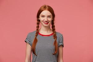 attrayant magnifique réconfortant jolie roux fille avec rouge lèvres, deux tresses, charmant sourire, spectacles blanc en bonne santé dents, habillé dans dépouillé T-shirt, isolé sur une rose Contexte photo