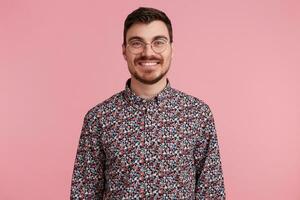 portrait de une agréable attrayant Beau Jeune homme portant des lunettes avec foncé cheveux non rasé avec barbe et moustache dans coloré chemise agréablement souriant, isolé plus de rose Contexte photo