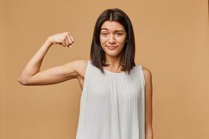 portrait de une fille sur de soi fier de sa force serre une poing et courbé sa bras à spectacle sa muscles, isolé plus de beige Contexte photo