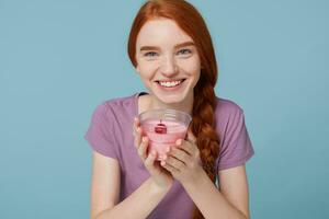 proche en haut de une content roux fille avec tresser sourit joliment détient une verre de Cerise yaourt dans sa mains, les usages laitier des produits isolé sur une bleu Contexte photo