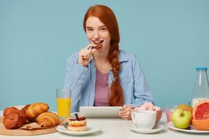 mignonne attrayant roux fille avec une sourire en mangeant biscuits séance à une tableau, dans sa mains en portant une tablette sur lequel lit le nouvelles, regards dans le caméra et sourires, contre une bleu Contexte. photo