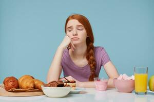 intérieur photo de roux fille à la recherche avec tristesse mécontentement sur cuisson des produits, pense à propos régime, supplémentaire calories, frais fruit jus yaourt sur le autre côté de tableau, isolé sur une bleu Contexte