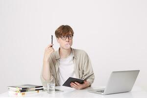 portrait de pensif Jeune homme étudiant porte beige chemise et des lunettes en pensant et soufflant bulles avec mastication gencive à le table avec portable ordinateur et des cahiers isolé plus de blanc Contexte photo