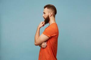 photo de une Jeune attrayant fort homme avec une à la mode la Coupe de cheveux épais barbe permanent de côté caméra habillé dans une rouge T-shirt détient le sien main près le sien menton reflète sur quelque chose pense philosophe.