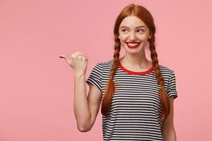 content excité roux fille avec deux tresses montrer du doigt avec pouce à le la gauche côté sur Vide copie espace et regards là avec intérêt, souriant largement, dessine attention, avec rouge lèvres, sur rose mur photo