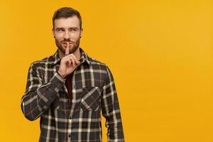 portrait de ruse, adulte Masculin avec brunette cheveux et poil. portant à carreaux chemise et accessoires. spectacles silence signe. en train de regarder à le droite à copie espace, isolé plus de Jaune Contexte photo