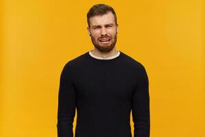 barbu déprimé gars, malheureux à la recherche homme avec brunette cheveux. a perçant. portant noir chandail. pleurs et tordu visage plein de larmes. en train de regarder à le caméra, isolé plus de Jaune Contexte photo