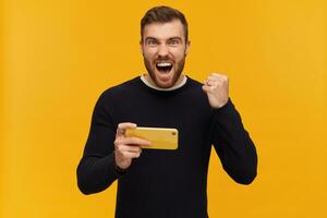 barbu gars, excité homme avec brunette cheveux. a perçant. portant noir chandail. en portant téléphone intelligent. serrer poing dans fête de la victoire. en train de regarder à le caméra, isolé plus de Jaune Contexte photo