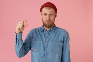 barbu Masculin dans denim chemise, renfrogné visage fait du une main geste, démontre quelque chose très minuscule, pose contre rose mur. une homme des offres à boisson une petit montant de liquide, à dépenser ensemble certains temps. photo