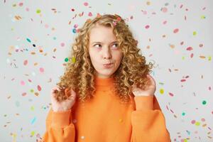 horizontal coup de attrayant Jeune femelle avec pensif contemplant expression, réfléchit à propos quelque chose, garde touche sa blond cheveux, habillés dans décontractée Orange chandail, des stands en dessous de chute confettis photo