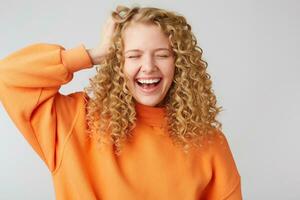 de bonne humeur insouciant joyeux aux cheveux bouclés blond détient sa main près tête fermé sa yeux de plaisir, des rires Heureusement, habillé dans une chaud Orange surdimensionné chandail, isolé sur blanc Contexte photo