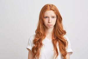 portrait de déprimé mignonne Jeune femme avec rouge ondulé longue cheveux et taches de rousseur porte t chemise se sent dérangé et offensé isolé plus de blanc Contexte photo