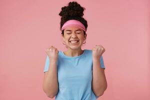 portrait de attrayant fille avec foncé frisé cheveux chignon. portant rose visière, des boucles d'oreilles et bleu T-shirt. a faire en haut. serrer sa les poings et plisser les yeux dans excitation. supporter isolé plus de pastel rose Contexte photo