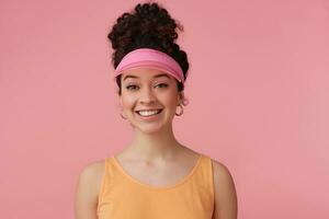 portrait de attractif, adulte fille avec foncé frisé cheveux chignon. portant rose visière, des boucles d'oreilles et Orange réservoir Haut. a faire en haut. en train de regarder avec sourire à le caméra isolé plus de pastel rose Contexte photo