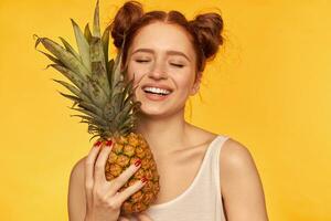 adolescent fille avec gros sourire et fermé yeux, content à la recherche rouge cheveux femme avec deux petits pains. portant blanc chemise et en portant ananas, en bonne santé mode de vie. supporter isolé plus de Jaune Contexte photo