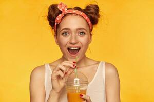 adolescent fille, content à la recherche rouge cheveux femme avec petits pains. portant blanc chemise et rouge adoré bandeau. à la recherche excité et en portant sa juteux frais. en train de regarder à le caméra isolé plus de Jaune Contexte photo