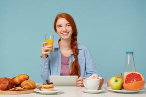 rêver à la recherche à le plus haut droite coin magnifique souriant roux fille en buvant Orange jus ayant petit déjeuner rougir nouvelles dans sa tablette, isolé sur bleu Contexte photo