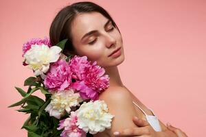 portrait de attractif, agréable à la recherche fille avec longue brunette cheveux, fermé yeux et en bonne santé peau. portant blanc robe et détient bouquet de fleurs. supporter isolé plus de pastel rose Contexte photo