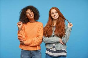 positif magnifique Jeune Dames à la recherche gaiement de côté et en jouant avec leur cheveux, étant dans agréable ambiance et souriant agréablement tandis que posant plus de bleu Contexte photo