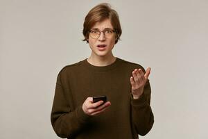 portrait de mécontentement, malheureux Masculin avec blond cheveux. portant marron chandail et lunettes. en portant une téléphone intelligent. confus de quoi il voit. en train de regarder renfrogné à le caméra isolé plus de gris Contexte photo