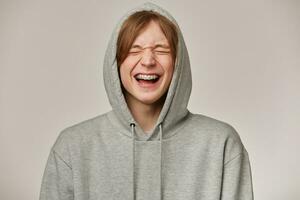 de bonne humeur homme, Beau gars avec blond cheveux. portant gris sweatshirt à capuche. a croisillons. gens et émotion concept. met capuche sur et en riant avec fermé yeux. supporter isolé plus de gris Contexte photo