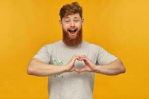 intérieur coup de Jeune barbu homme, avec rouge cheveux et gros barbe, spectacles cœur signe avec le sien mains, sourit largement et se sent content, isolé plus de Jaune Contexte. photo