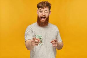 intérieur coup de Jeune Beau gingembre Masculin avec une gros barbe, montrer du doigt avec tous les deux mains dans caméra, sourit et clin d'œil. isolé plus de Jaune Contexte photo