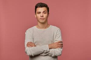 Jeune Beau malheureux gars porte dans à carreaux chemise, regards à le caméra avec dégoûté expression, croisé bras, isolé plus de rose Contexte. photo