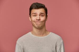 Beau Jeune triste gars avec élevé les sourcils porte dans Vide T-shirt, regards à le caméra avec méfiance, des stands plus de rose Contexte. photo