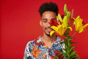 proche en haut de Jeune content africain américain homme, porte dans hawaïen chemise, regards à le caméra avec content expression, détient Jaune et rouge fleurs et couvert affronter, des stands plus de rouge Contexte. photo