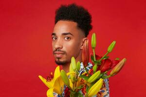 proche en haut de Jeune calme foncé écorché homme, porte dans hawaïen chemise, détient Jaune et rouge fleurs bouquet, des stands plus de rouge Contexte. photo