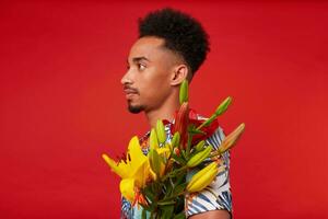 portrait de Jeune calme africain américain homme, porte dans hawaïen chemise, détient Jaune et rouge fleurs bouquet, des stands plus de rouge Contexte. photo