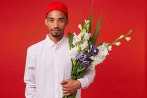 Jeune calme africain américain gars dans blanc chemise et rouge chapeau, détient bouquet , des stands plus de rouge Contexte et regards à le caméra. photo