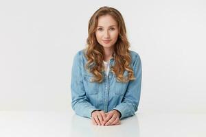 portrait de Jeune mignonne frisé blond Dame dans denim chemises séance à le blanc table et sourire, regards positif, isolé plus de blanc Contexte. photo