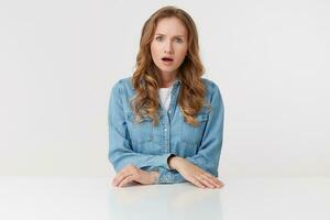 photo de Jeune indigné blond femme porte dans denim chemises, séance à le blanc tableau, renfrogné et regards mécontent, isolé plus de blanc Contexte.