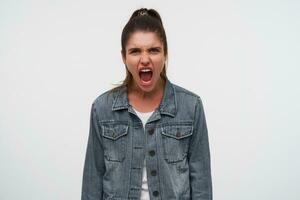 portrait de Jeune agressif brunette Dame porte dans blanc T-shirt et denim vestes, en hurlant et regards en colère, des stands plus de blanc Contexte. photo