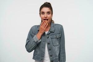 Jeune content étonné brunette Dame dans blanc T-shirt et denim vestes, regards à le caméra avec large ouvert bouche et yeux dans surpris expression, couvert bouche avec palmier, des stands plus de blanc Contexte. photo