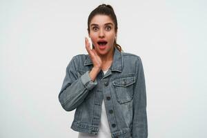 Jeune content étonné brunette Dame dans blanc T-shirt et denim vestes, regards à le caméra avec large ouvert bouche et yeux dans surpris expression, garder paume sur le joue, des stands plus de blanc Contexte. photo