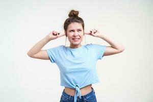 mécontent Jeune jolie brunette femelle avec chignon coiffure renfrogné sa visage et couvrant oreilles tandis que éviter bruyant des sons, permanent plus de blanc Contexte photo