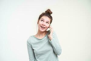 heureux Jeune attrayant aux yeux bleus brunette femelle à la recherche gaiement vers le haut tandis que ayant agréable téléphone conversation, isolé plus de blanc Contexte dans décontractée porter photo