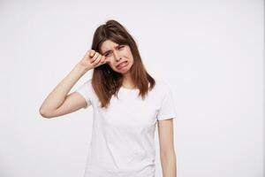 studio photo de morne Jeune brunette Dame avec décontractée coiffure essuyage larmes et renfrogné Malheureusement sa visage tandis que permanent plus de blanc Contexte dans blanc de base T-shirt