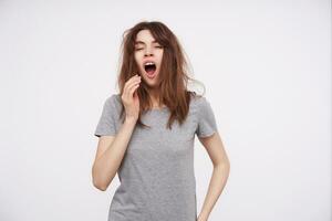 studio photo de Jeune somnolent marron aux cheveux femelle habillé dans de base gris T-shirt élevage main à sa bouche tandis que bâillement avec fermé yeux, isolé plus de blanc Contexte