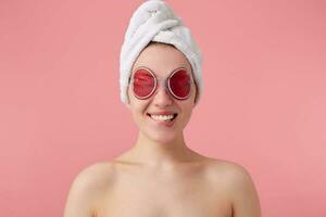 proche en haut de content Jeune femme après spa avec une serviette sur sa diriger, avec masque pour yeux, largement sourires, regards positif, des stands plus de rose Contexte. photo