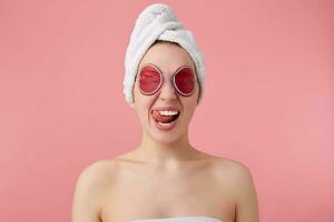 proche en haut de Jeune marrant Dame avec masque sur yeux, après douche avec une serviette sur sa diriger, regards à le caméra, spectacles langue et des stands plus de rose Contexte. photo